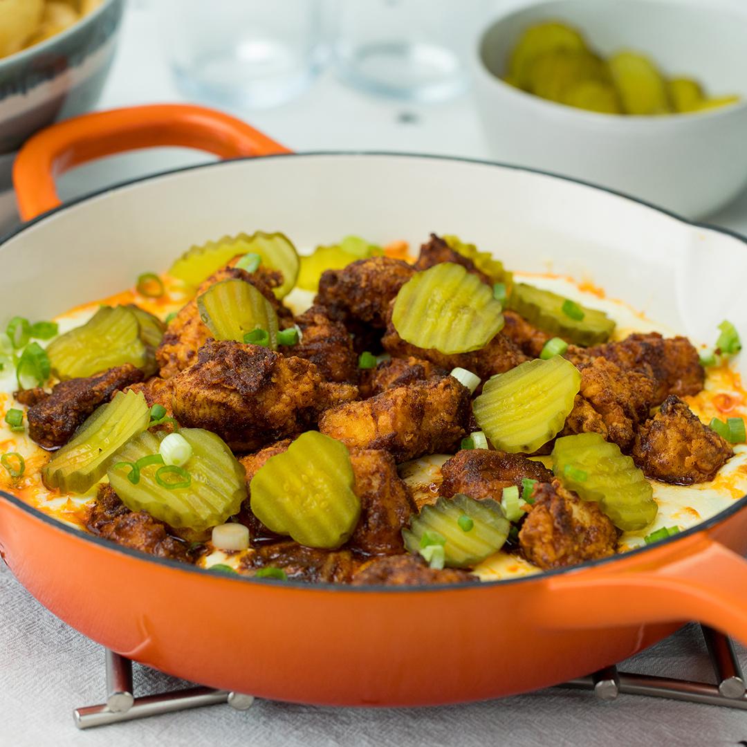 A dish of fried chicken pieces topped with pickles and green onions in a pan, served with a bowl of pickles in the background