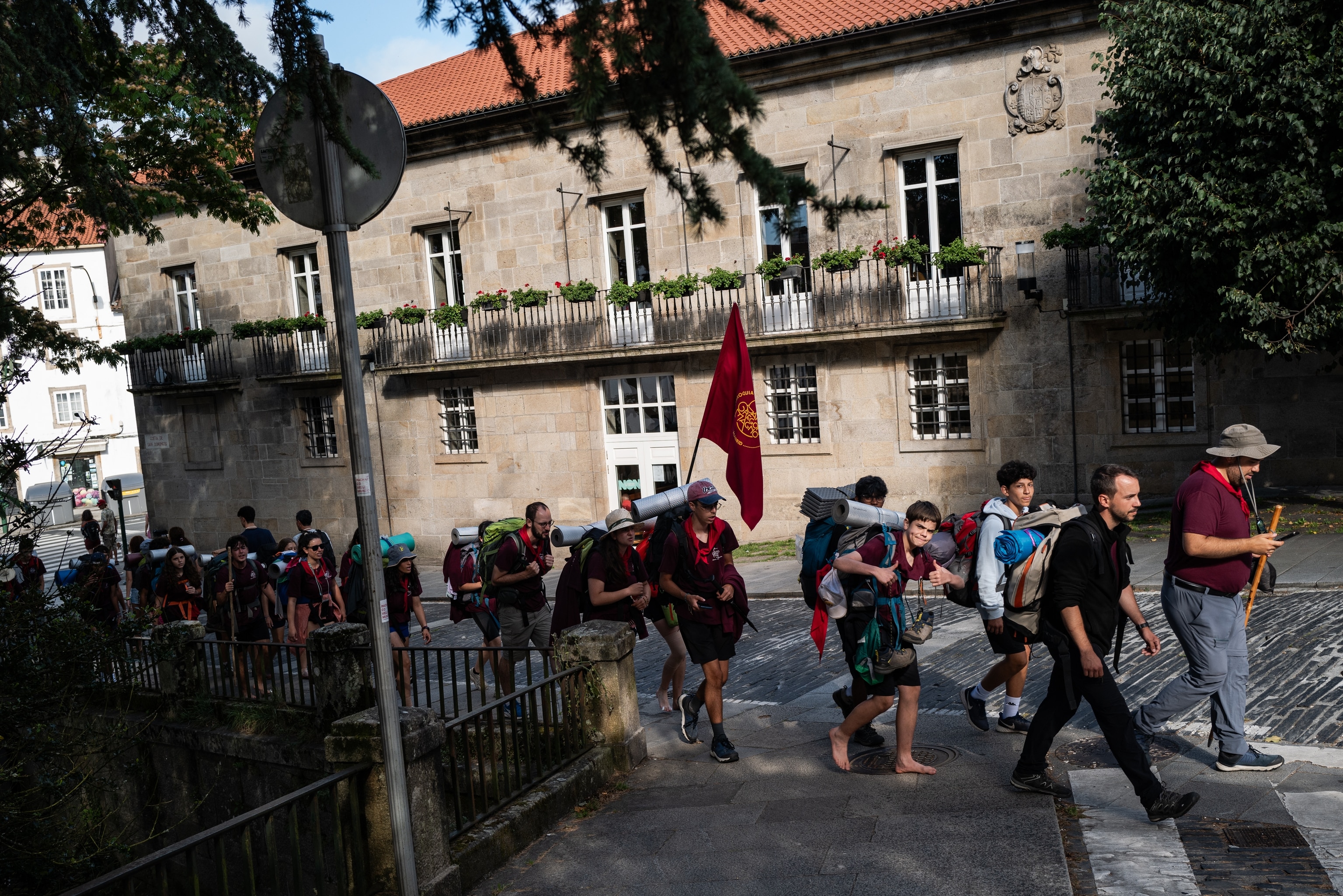 A group of hikers wearing backpacks on a shady street in small-town Spain. One carries a flag and some are barefoot.