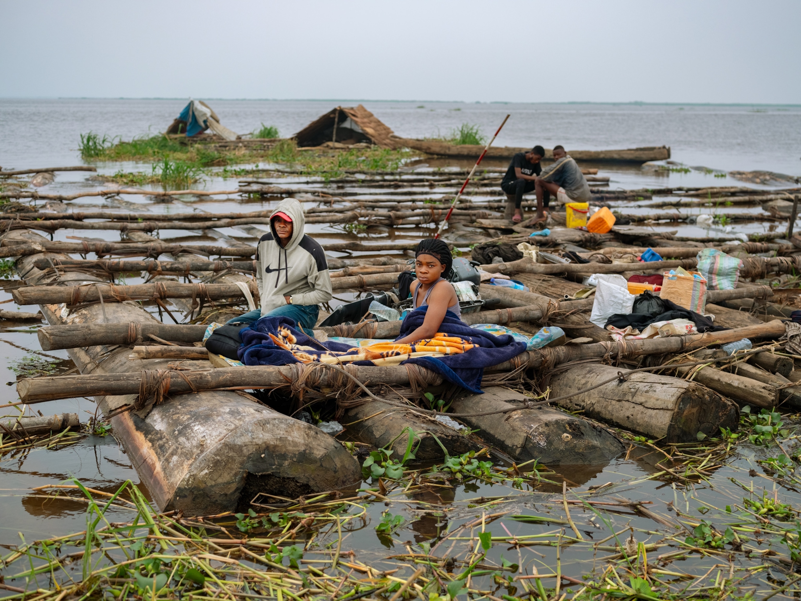 Two individuals sit atop a raft made of logs with their belongings around them.
