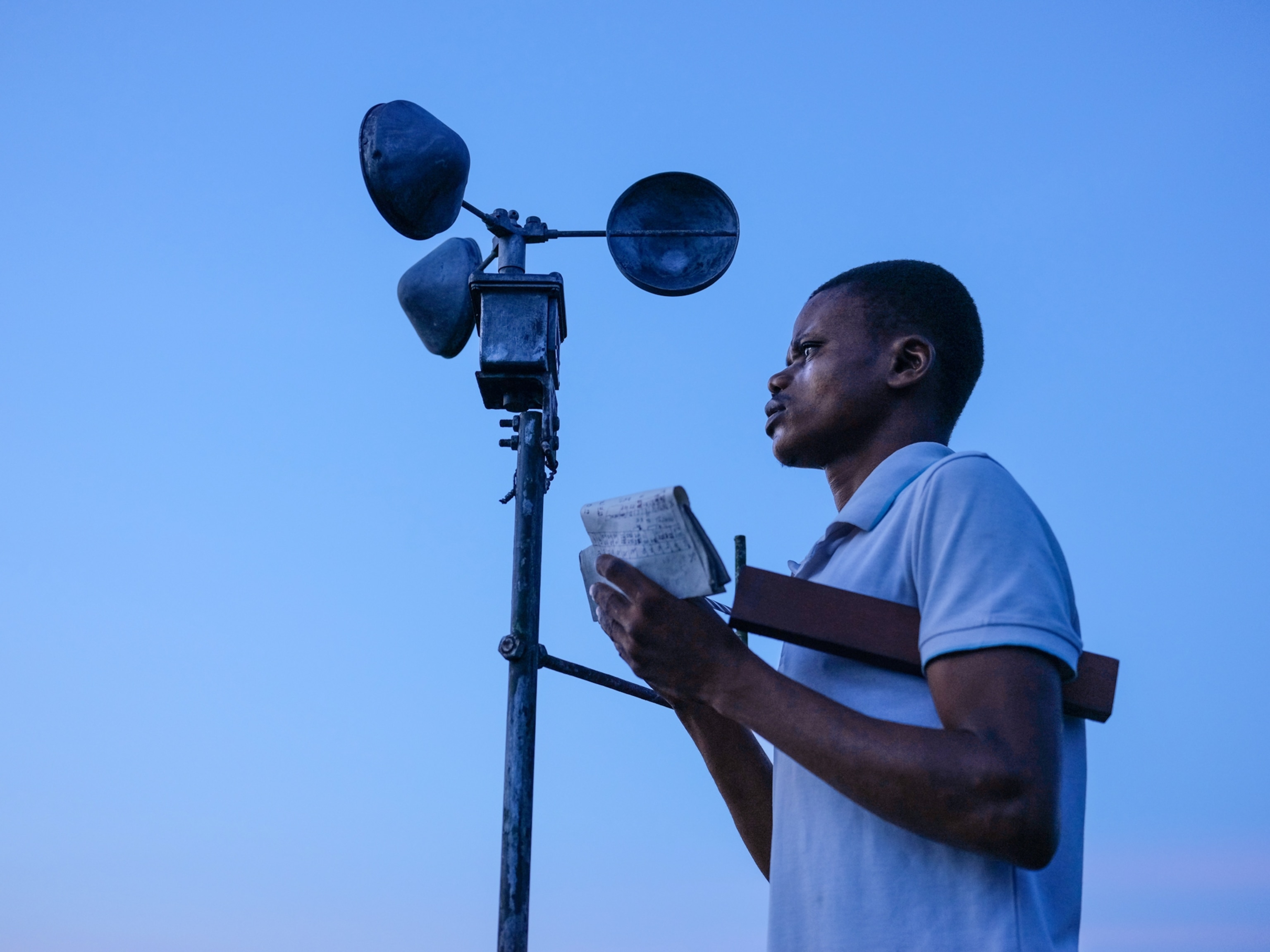Michel Atianga Bongombe is pictured in front of a tall weather instrument, holding paper and a pen.