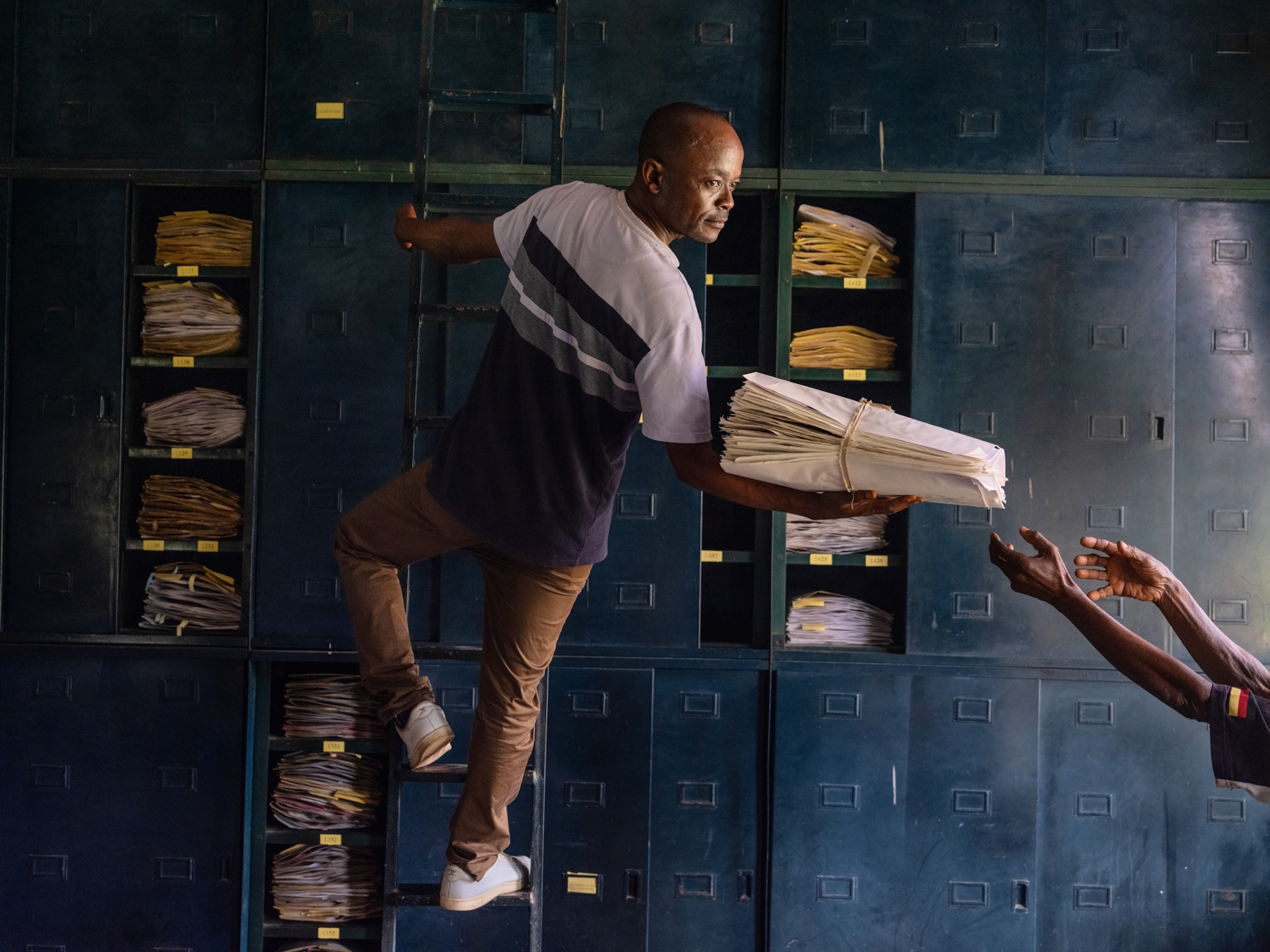 Archivist Fidèle Noa Manganga is standing on a ladder, holding onto it with his left hand. He is turned to the right handing white packages of dried specimen to another employee.