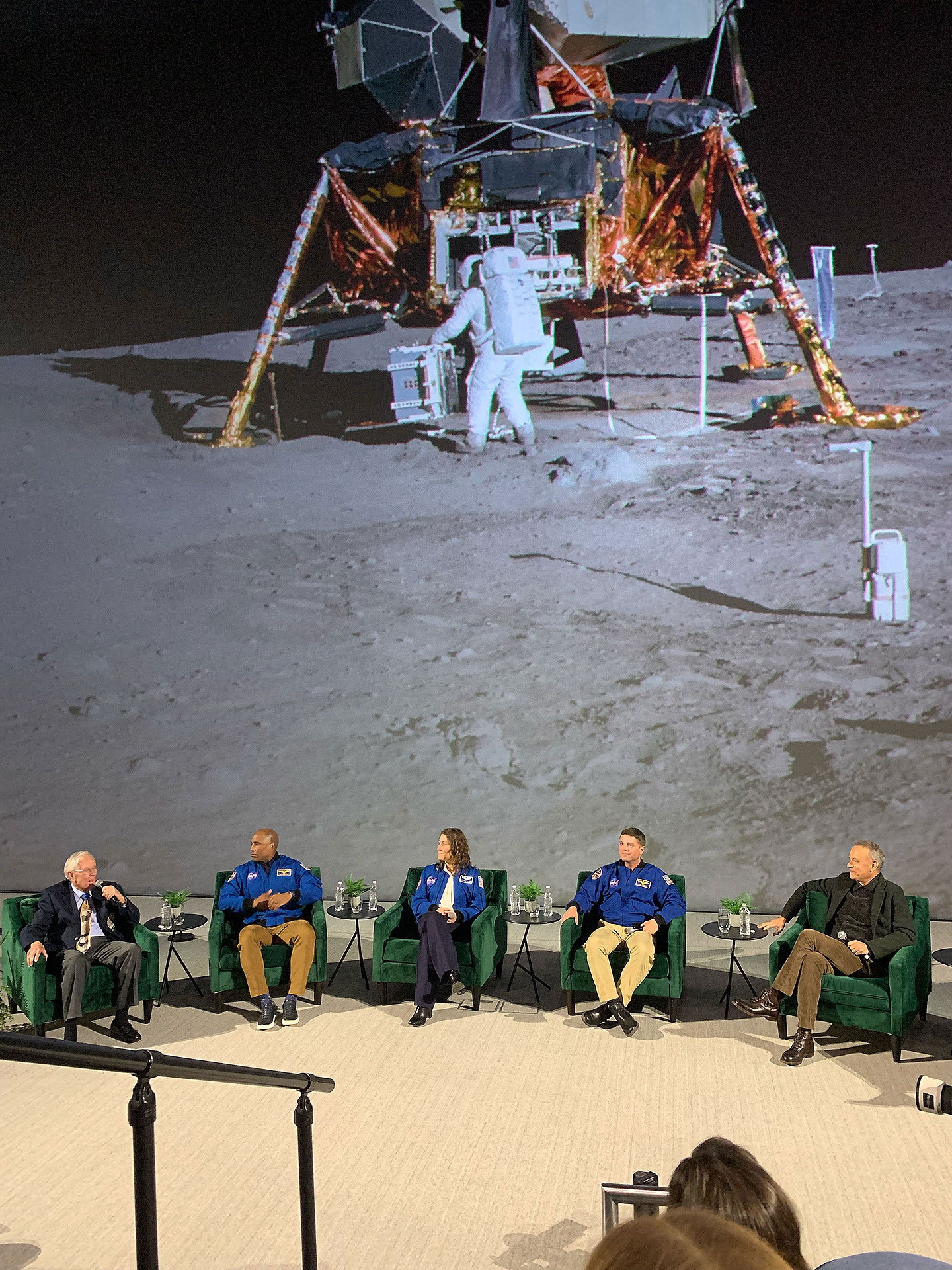 a panel discussion with astronauts and actor Tom Hanks at the base of a 5-story-tall theater screen showing a moon lander and astronaut