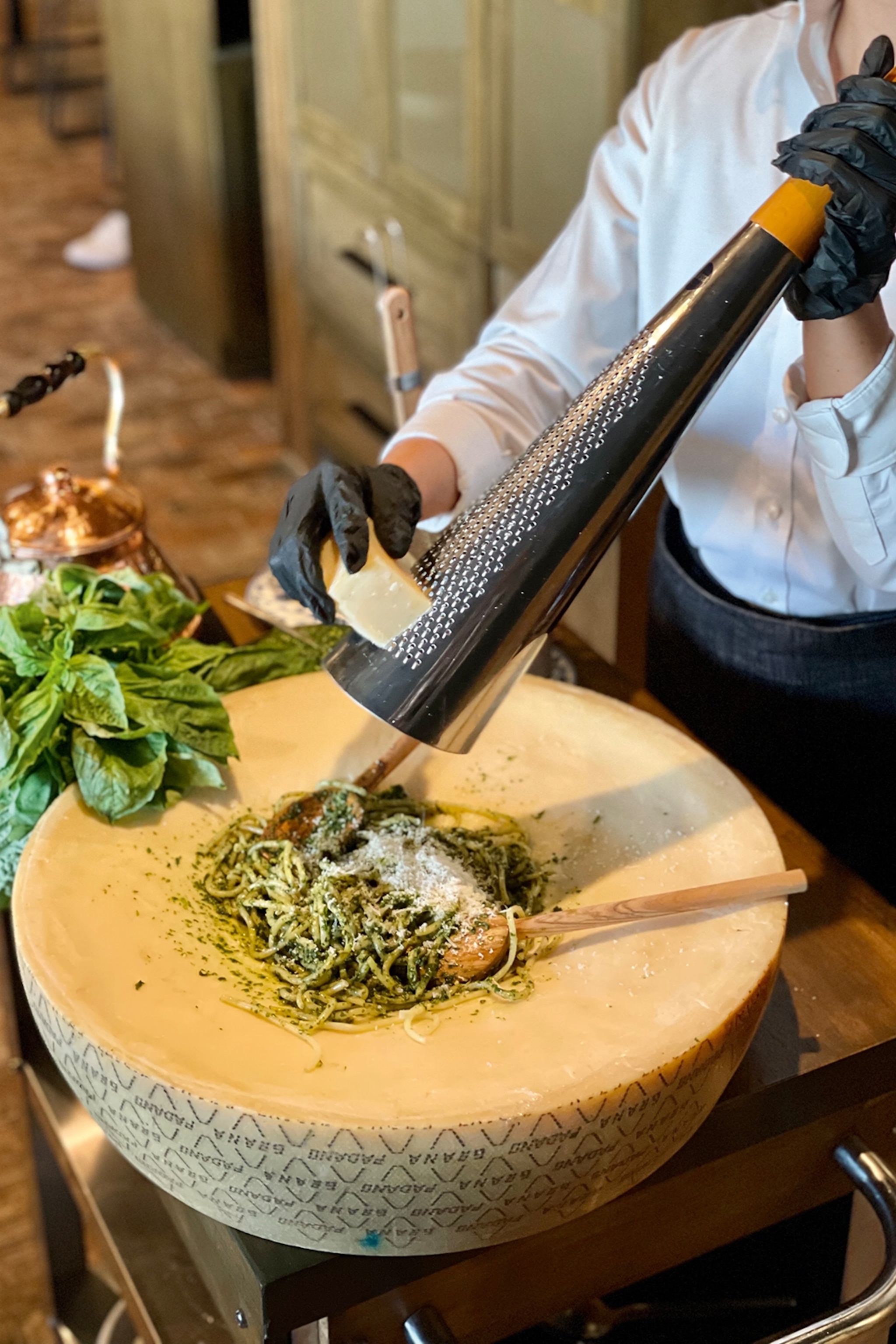 A dynamic shot of a gloved waiter adding parmesan cheese on a portion of pesto pasta in a cheese wheel.