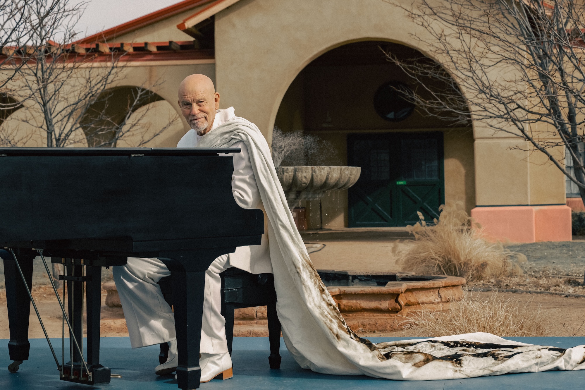John Malkovich sits at a piano and smirks.