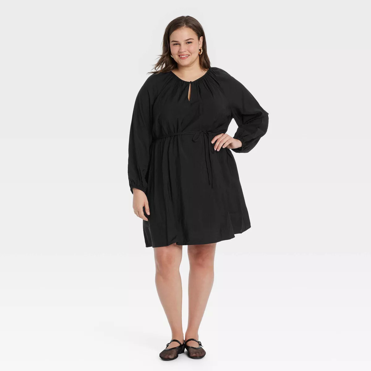 Person in a casual, long-sleeved black dress poses with one hand on hip, wearing flats and earrings, smiling