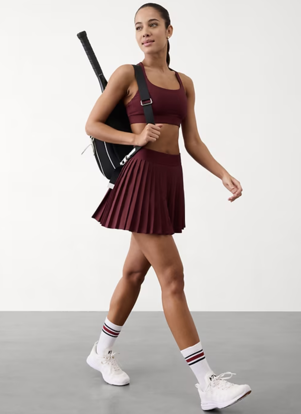 Model in athletic crop top and pleated skirt, carrying a tennis racket on her back, wearing sporty shoes and striped socks