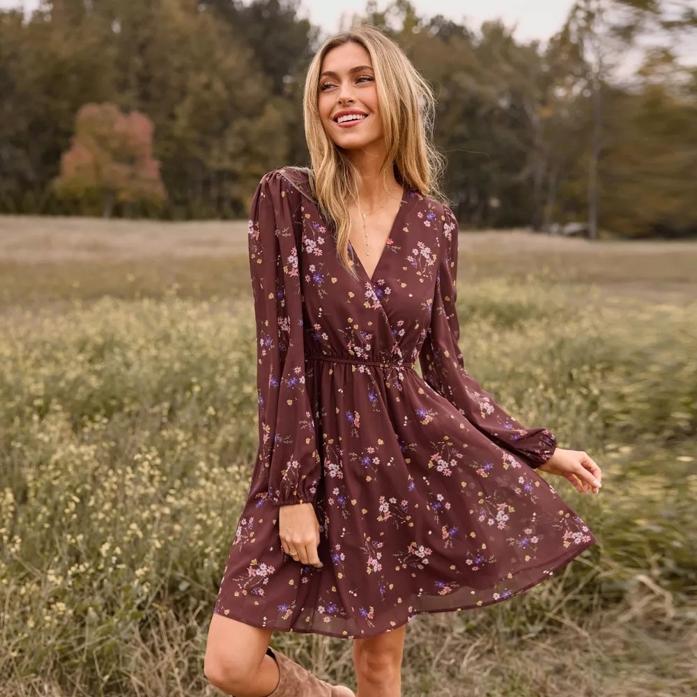 Person wearing a burgundy floral-patterned dress in a field, smiling. The dress is knee-length with long sleeves and a cinched waist