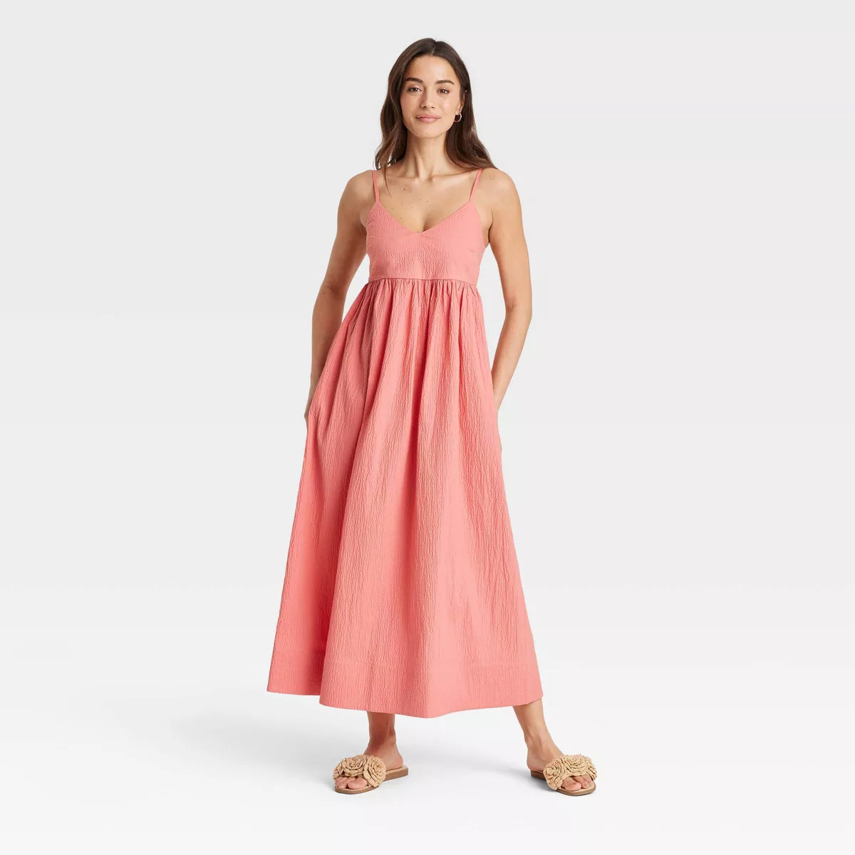 Person wearing a sleeveless, flowing dress poses with hands in pockets, paired with woven sandals, styled for a casual setting
