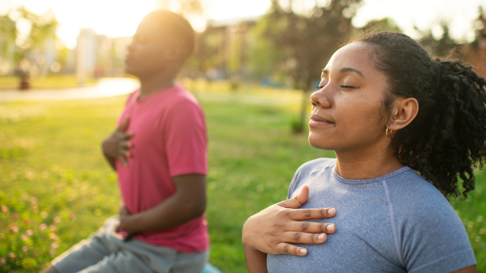 a man and a woman practice yoga outside, resting with their hands on their hearts and eyes closed as if taking a deep breath