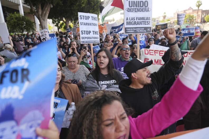 A crowd of people march while holding up signs and raising their fists criticizing immigration raids.
