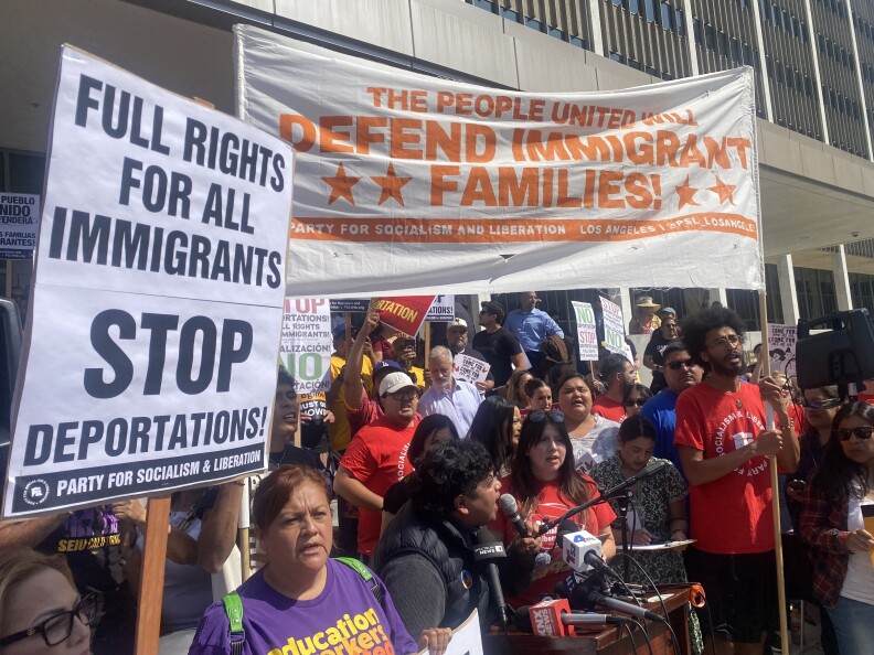 Dozens of people attend a rally/ news conference in downtown Los Angeles. One man with dark hair and brown skin appears to be speaking into a microphone. Other people around him hold signs and banners. One banner reads: