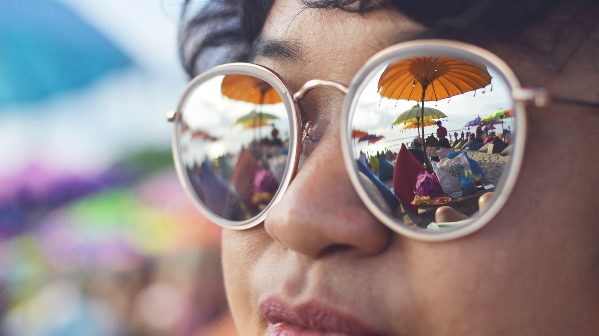 Horizontal close up shot of a girl with reflective sunglasses, mirroring a colorful beach club. Messy hair, Asian ethnicity.