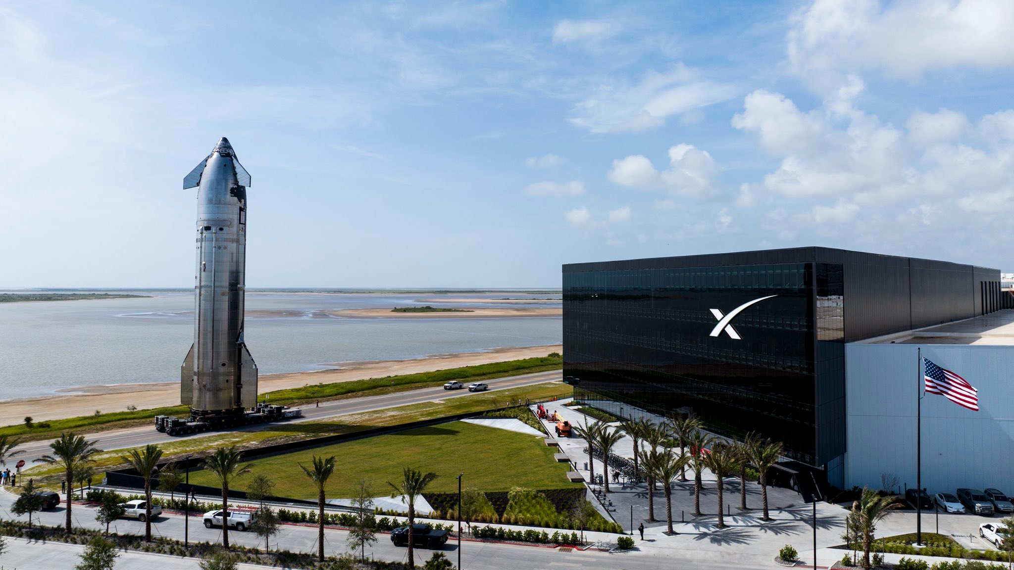 a large silver spacecraft rolls down a seaside road next to a large black building