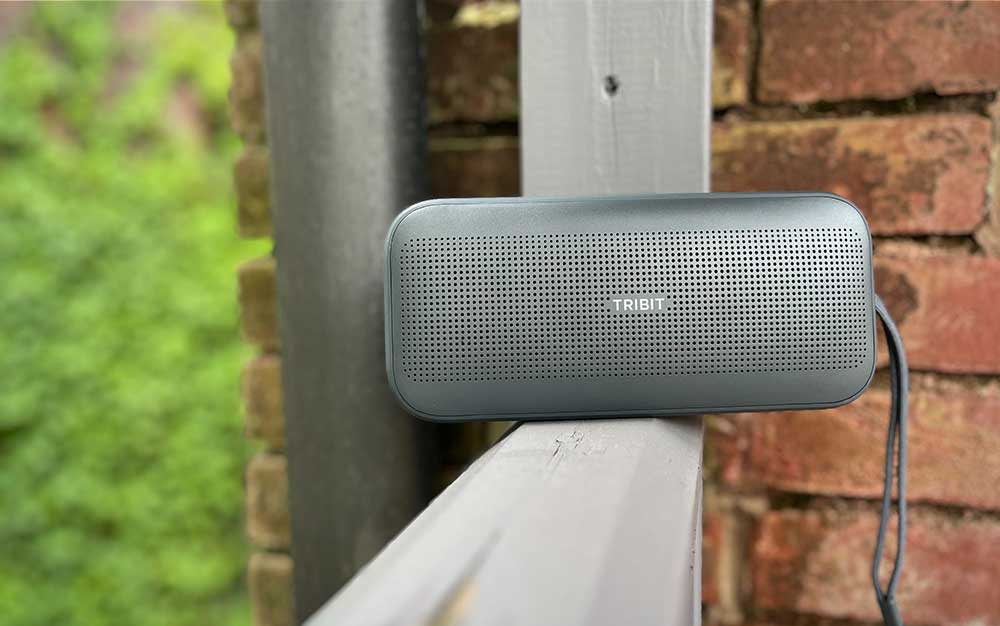 A gray Tribit StormBox Flow portable speaker on a wooden railing outdoors.