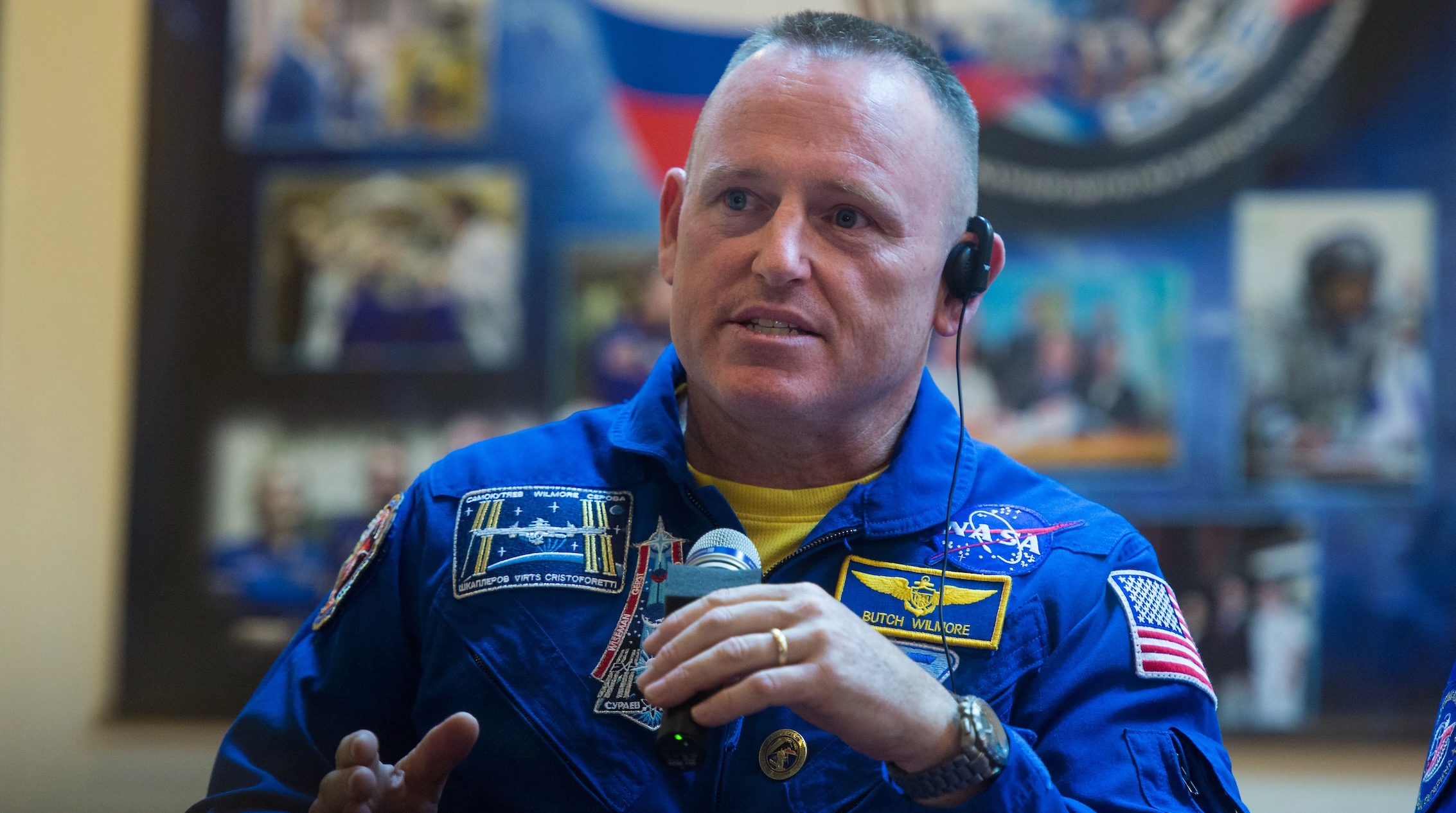 a middle-aged astronaut with a buzz cut and wearing a blue flight suit speaks into a microphone
