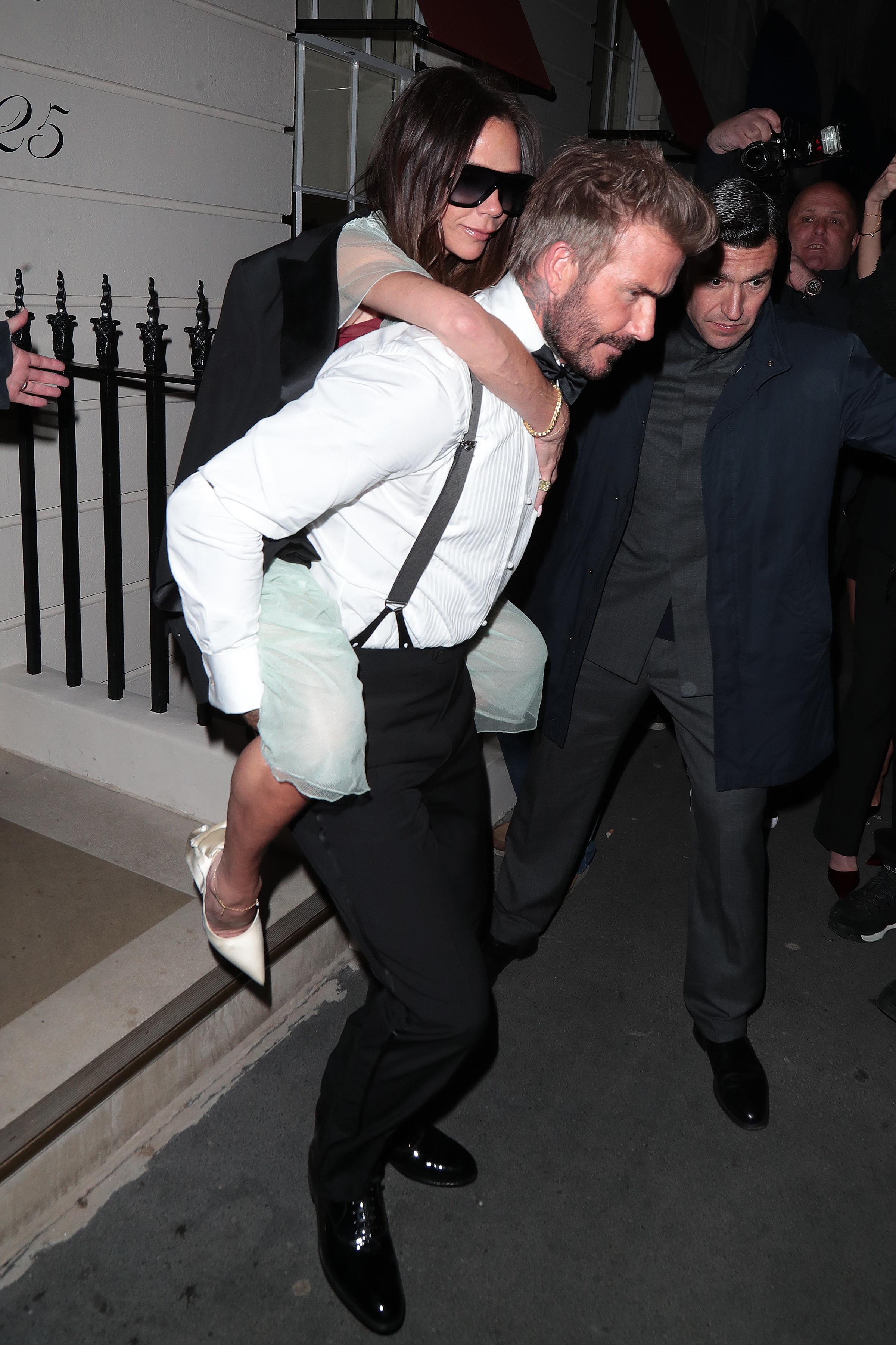 David Beckham in a white shirt, suspenders and black pants, carries out Victoria Beckham in a blue dress and black jacket.