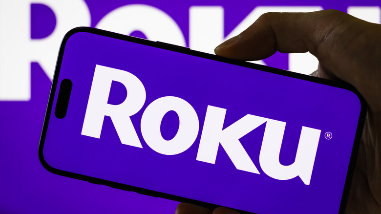 A person holding a phone that displays the Roku logo, set against a backdrop with that is also the Roku logo.