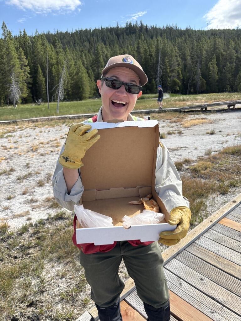 Yellowstone National Park Geology Program volunteer Tara Cross smiles and shows off a pizza box found on Geyser Hill in Upper Geyser Basin. CREDIT: National Park Service photo by Margery Price, August 2025.