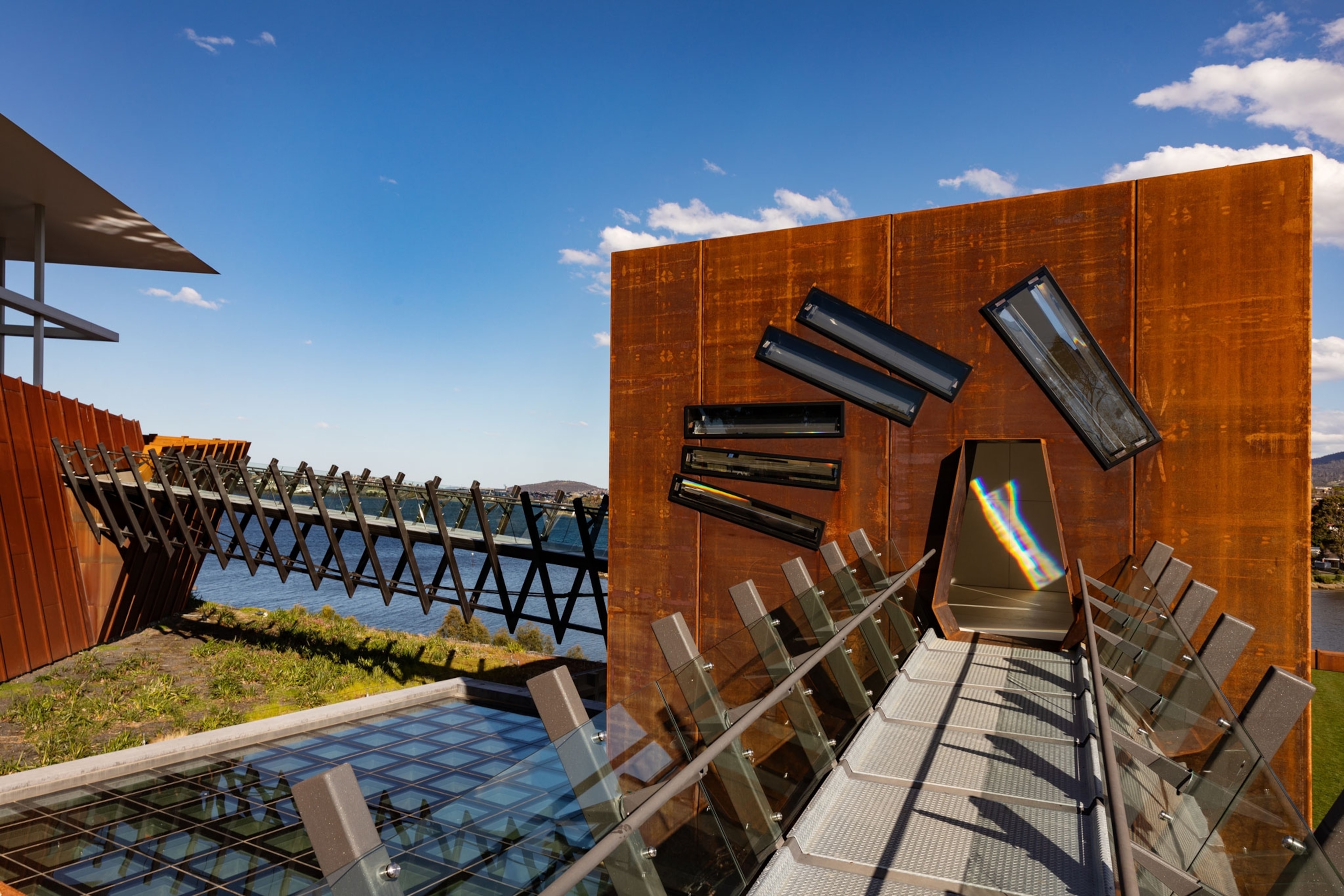 A modern architectural structure featuring a glass walkway leading to a rust-colored steel building with angled windows, overlooking a body of water under a clear blue sky.