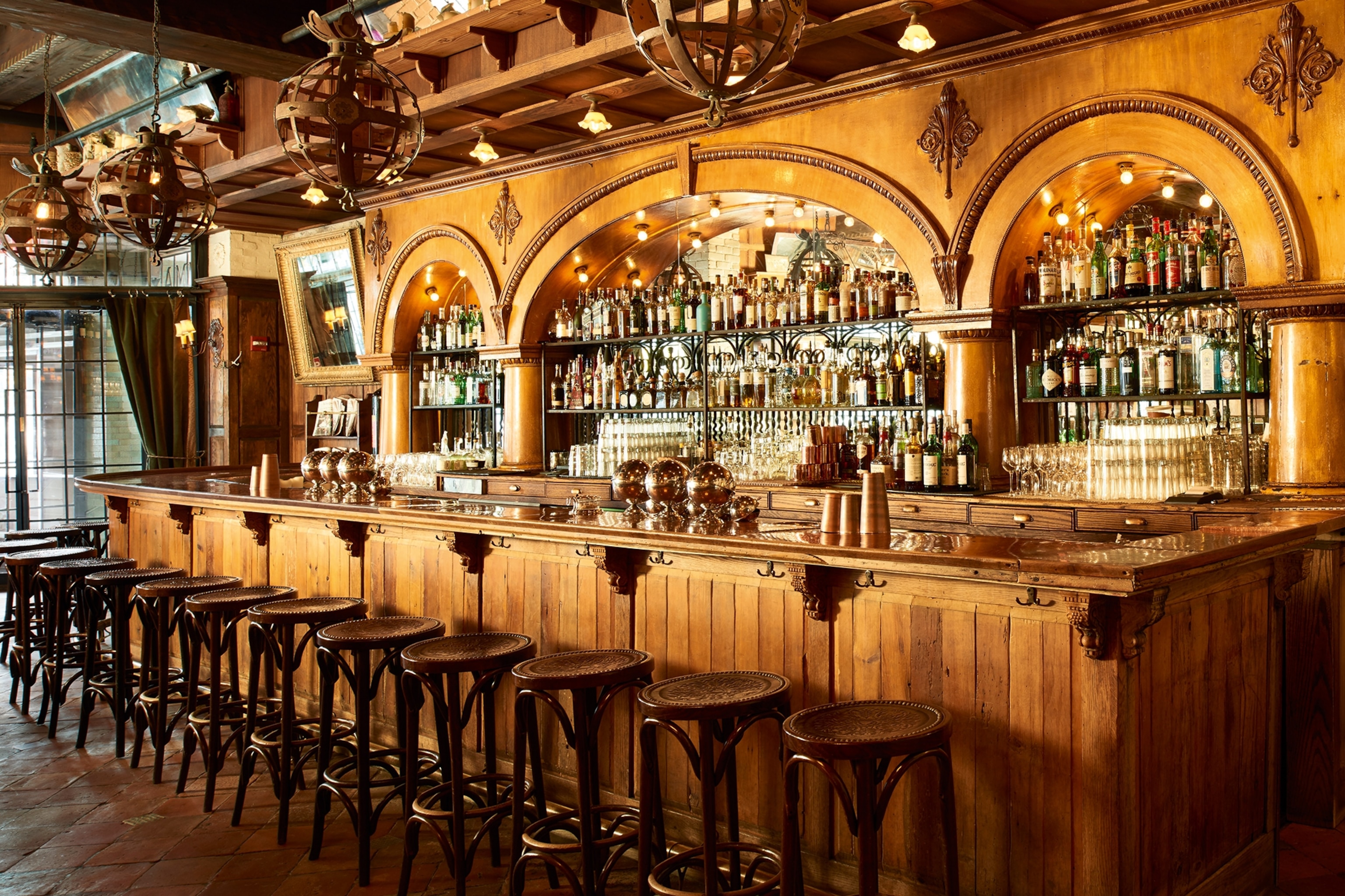 A landscape shot of a warm, wooden and brass restaurant bar with old-school stools, globe-caged lamps and arched alcohol wall cases behind the counter.