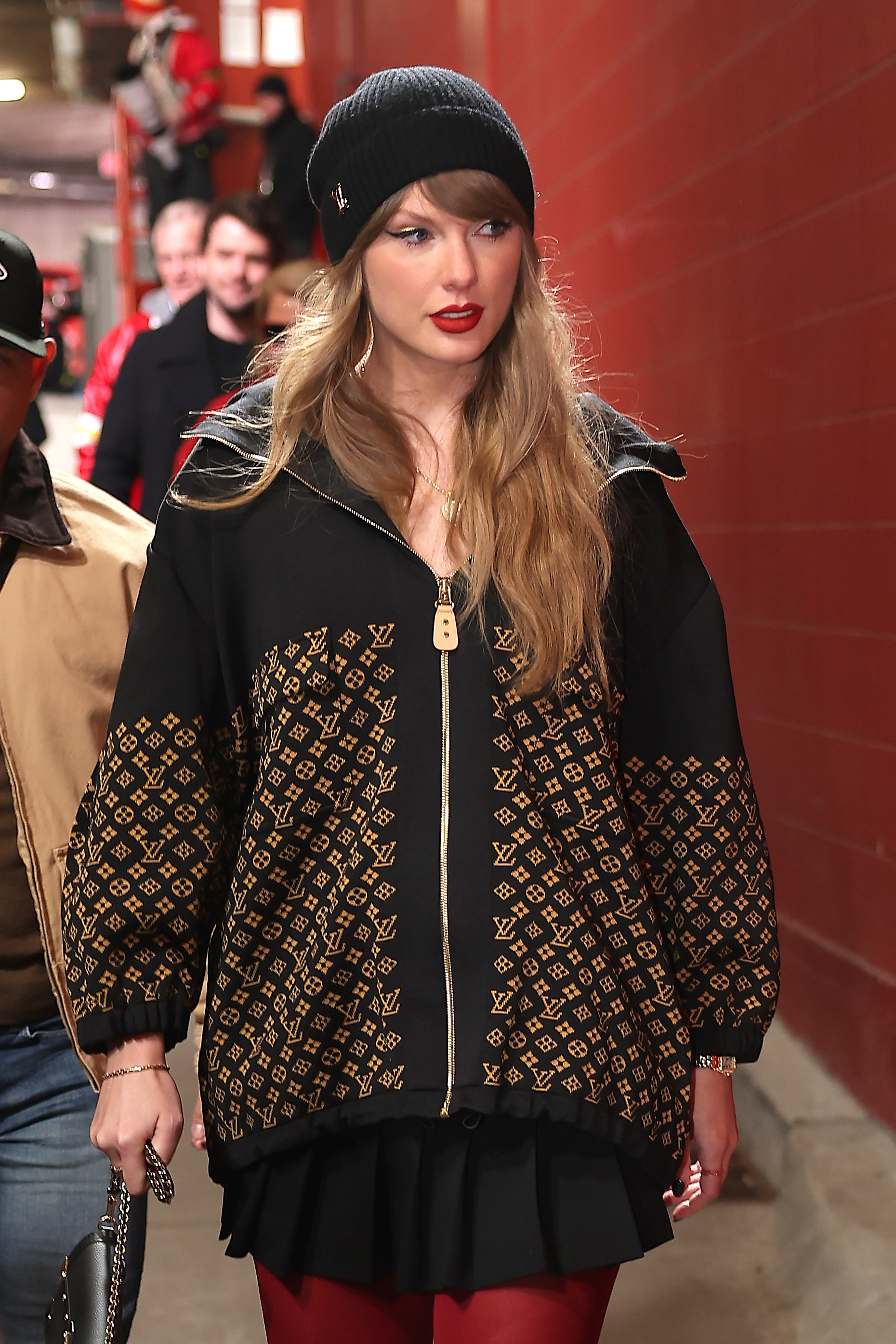 Taylor Swift wears a black beanie, black Louis Vuitton zip-up jacket, black skirt, and red tights.