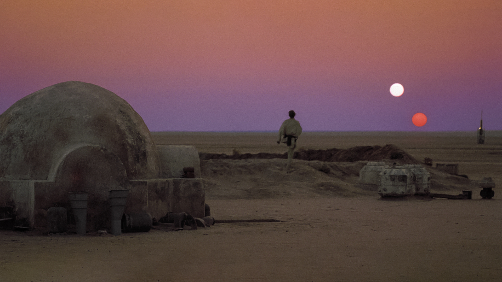 A man wearing white stands outside a stone igloo in the middle of a desert with a red and white sun in the dusky red sky.
