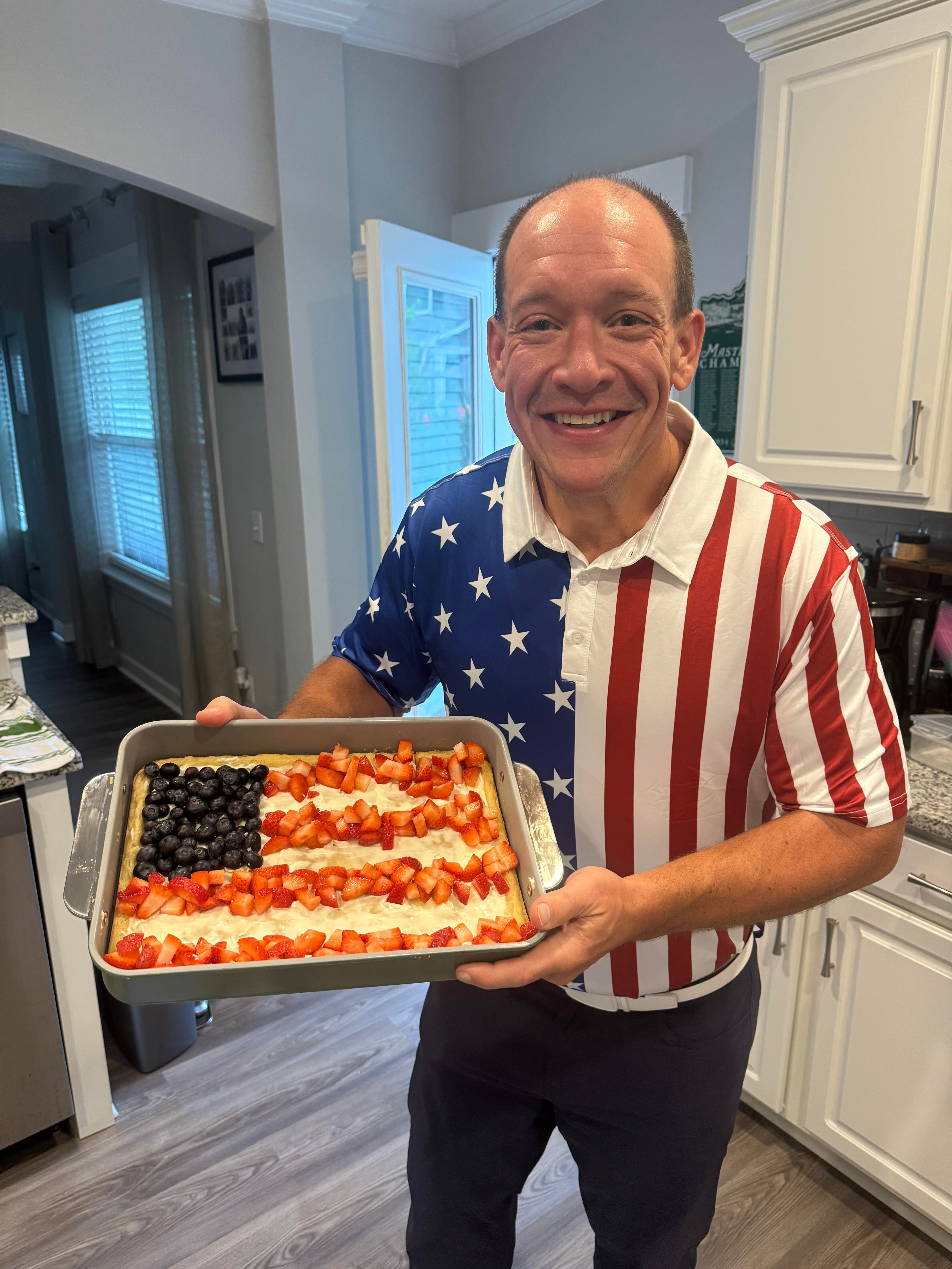 smiling man in a kitchen with a red, white, blue shirt holding an decorated fruit tart tray in the colors of the american flag